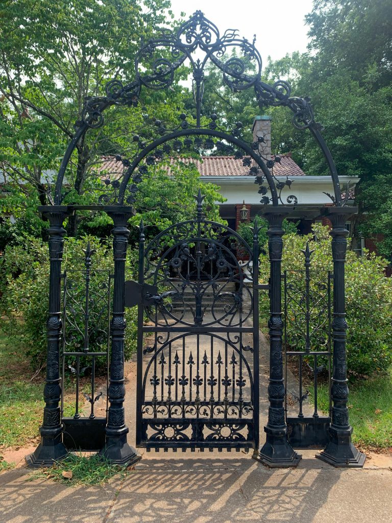 Classic wrought iron gate with intricate design in front of traditional house, surrounded by greenery.